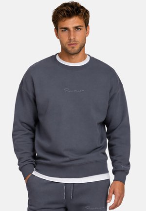 Uomo che indossa una felpa grigio scuro e pantaloni abbinati con sottili loghi testuali, in piedi con le mani nelle tasche davanti a uno sfondo semplice.