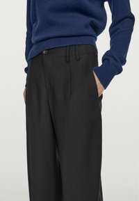 Zwarte op maat gemaakte broek met zijzakken, plooien en een gladde stofafwerking. Gecombineerd met een blauwe gebreide trui, wat de contrast in textuur benadrukt.