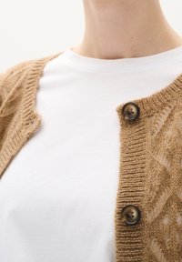 Beige gebreid vest met een gestructureerd, open patroon. Voorzien van donkere knopen en een witte T-shirt met ronde hals eronder.