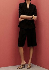 Czarna marynarka i pasujące szorty sięgające kolan, wykonane z teksturowanej tkaniny, połączone z wysokimi mule w wężowy wzór. Różowe tło.