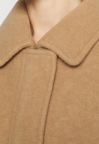 Wollmantel in Kamelhaarfarbe mit weitem Kragen, sichtbarer Textur und einem klaren, minimalistischen Design mit verdecktem Verschluss.