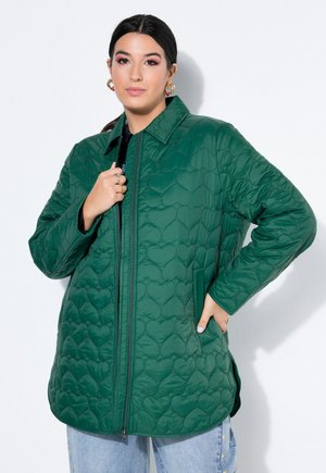 Kvinde iført en grøn quiltet jakke med hjerte-mønstre og lyseblå jeans, stående med den ene hånd i lommen og den anden nær kraven.