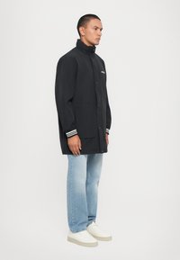 Black lightweight jacket with a high collar, button front, and striped cuffs, paired with light blue jeans and white sneakers.