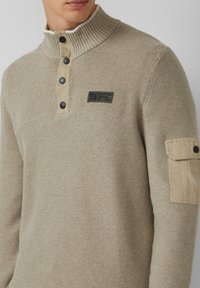 Beżowy sweter o teksturowanym splotach, z żebrowanym kołnierzem typu mock, czterema czarnymi guzikami, naszywką na klatce piersiowej z napisem "Elevated Casual Est. 1969" oraz kieszonką na rękawie.