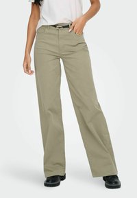 Femme portant un pantalon beige à jambes larges, une ceinture noire, un haut blanc rentré, et des chaussures noires épaisses, debout avec une main dans la poche.