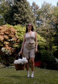Bežs svītru crop tops un šortu komplekts, tur rokās vītņu grozu, kas piepildīts ar baltām margrietiņām. Tajā ir arī saulesbrilles un baltas potītes zeķes ar mežģīņu apdari.