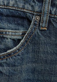 Denimová látka v tmavomodrej farbe s viditeľným prešívaním v svetlohnedej farbe. Detailný záber na prednú vrecko a kovový rýfkový detail. Mäkka textúra.