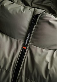 Nærbillede af en olivengrøn quilted jakke lynlås med sort trækstrop, der har hvid "SUPERDRY" branding og en lille orange rektangulær detalje.