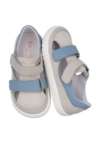 Zapatos infantiles de cuero gris y azul con correas de velcro, lados abiertos y una superficie texturizada en el tejido gris. Suela de goma blanca.