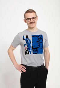 Šedé tričko s modrou grafikou kraví hlavy a textem "LA RACHE UNI RIT" v černé barvě. Nošeno s černými kalhotami.