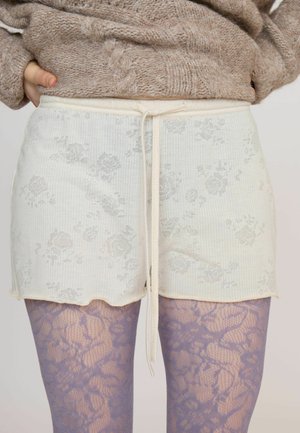 Personne portant un short en tricot beige à motif floral avec un cordon et des collants violets à motif dentelle, associés à un gros pull en tricot marron.