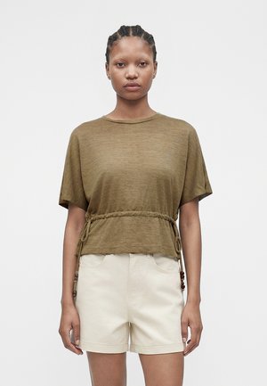 Vrouw met gevlochten haar die een olijfgroene top met korte mouwen en een strakke taille en off-white korte broek draagt, staand tegen een effen achtergrond.