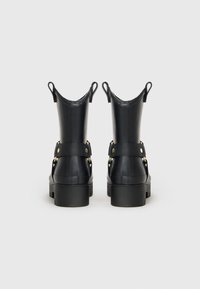 Ζεύγος μαύρων ankle boots με χοντρές σόλες, με λαβές στο πάνω μέρος και διακοσμητικές ζώνες με χρυσές λεπτομέρειες, εμφανίζεται από πίσω σε απλό φόντο.
