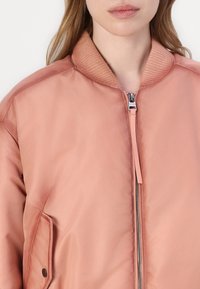 Padded bomber jacket in soft pink fabric. Features a ribbed collar, front zipper, and side pocket with snap button. Smooth texture.