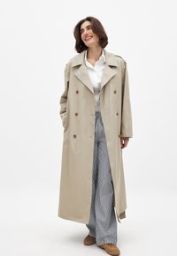Beige trench coat dengan desain double-breasted, kerah besar, dan saku samping, dipadukan dengan kemeja putih dan celana lebar bergaris.