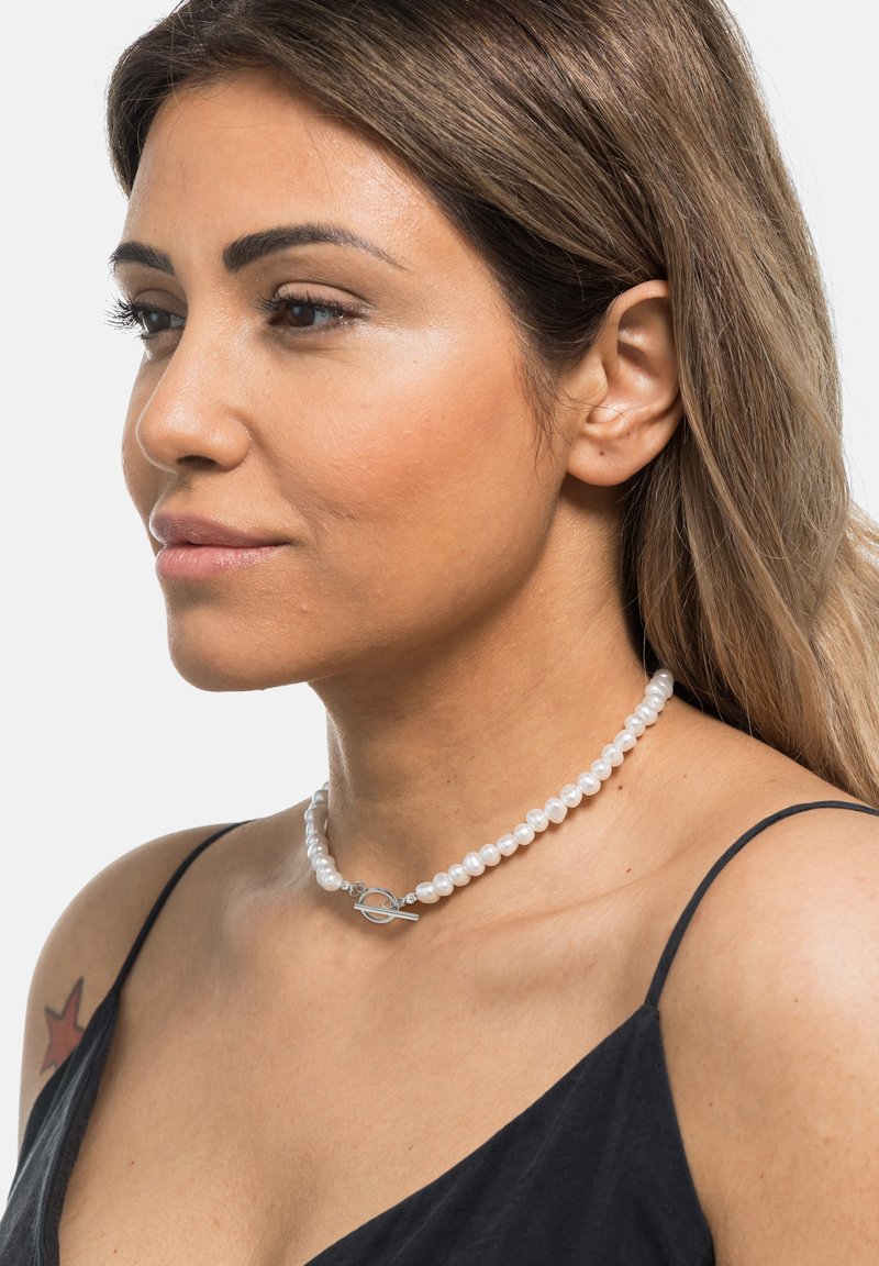 Donna con capelli castano chiaro che indossa una collana di perle con chiusura a toggle in argento e un top nero senza maniche, rivolta verso sinistra su sfondo bianco.