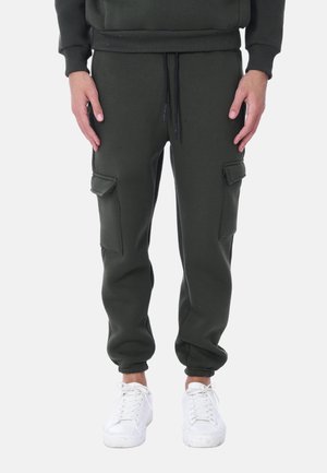 Pantaloni jogger verde scuro con polsini elastici, due tasche cargo, vita con coulisse e tessuto morbido e strutturato. Abbinati a sneakers bianche.