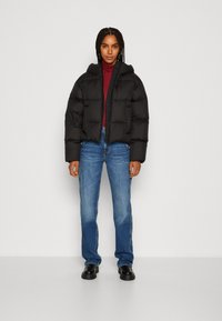 Schwarze Steppjacke mit Kapuze, mit einer kastigen Passform und gestepptem Textil; getragen über einem kastanienbraunen Rollkragenpullover und blauen Jeans. Schwarze Schuhe.
