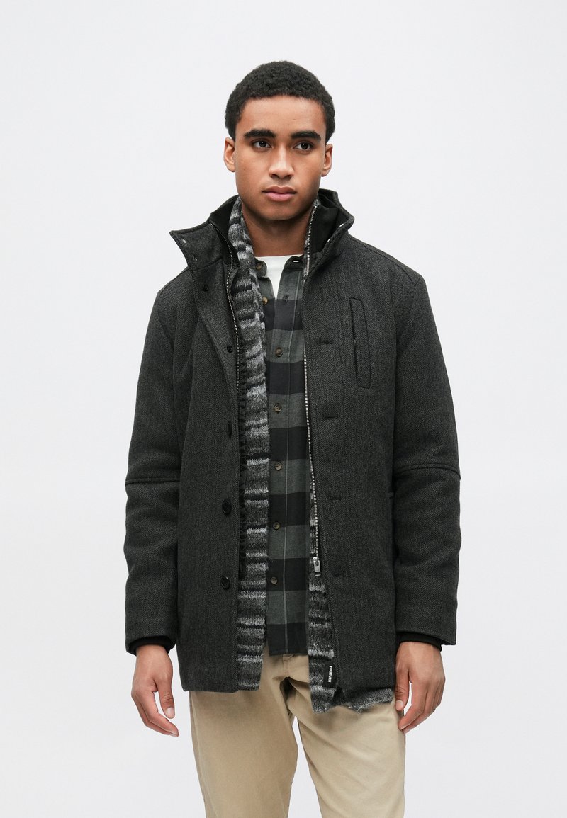 Casaco cinzento em mistura de lã com gola alta, fecho de botões e bolsos frontais. Usado por cima de uma camisa às riscas com um cachecol texturado e riscado.