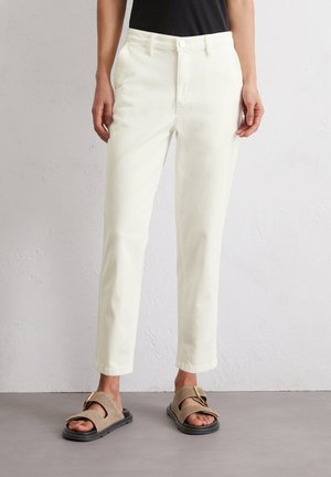 Person, die hoch taillierte, gerade geschnittene, cremeweiße Hose, schwarzes Oberteil und beige Sandalen mit dicken Riemen und goldenen Schnallen trägt, steht auf grauem Boden.
