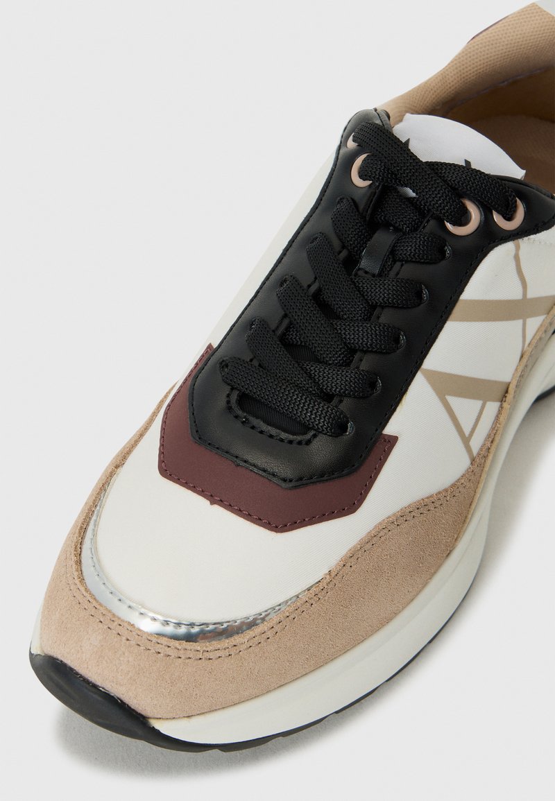 Zapatillas de deporte con cordones que presentan una combinación de materiales: parte superior sintética negra, acentos de ante beige, cuerpo blanco y detalles metálicos.