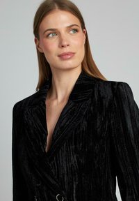 Blazer doppiopetto in velluto nero con revers pronunciati, texture a coste e bottoni circolari argentati, che mette in risalto una silhouette aderente.