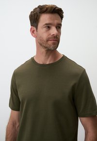 Maglietta verde oliva con scollo rotondo, maniche corte e una texture liscia. Il tessuto sembra leggero e informale.