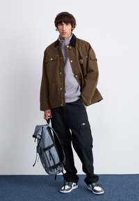 Smeđi praktični kaput s zlatnim gumbima, sivi zip-up pulover, crne cargo hlače i siva ruksak. Crno-bijeli tenisice upotpunjuju izgled.