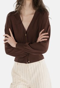 Brązowy kardigan z dekoltem w serek, zapinany na guziki, z prążkowanymi mankietami, w połączeniu z beżowymi spodniami w paski. Gładki materiał, luźny krój.