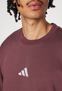 Bordeaux sweatshirt met een ronde halslijn en een wit logo met drie diagonale strepen op de borst. Stof lijkt zacht en textuur te hebben.