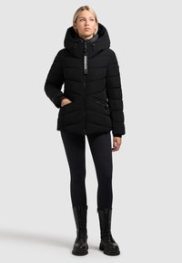 Jachetă puffer neagră cu glugă largă, închidere cu fermoar și cusături orizontale. Dispune de buzunare cu fermoar și un eticheta cu logo. Purtaa cu leggings negri și cizme.
