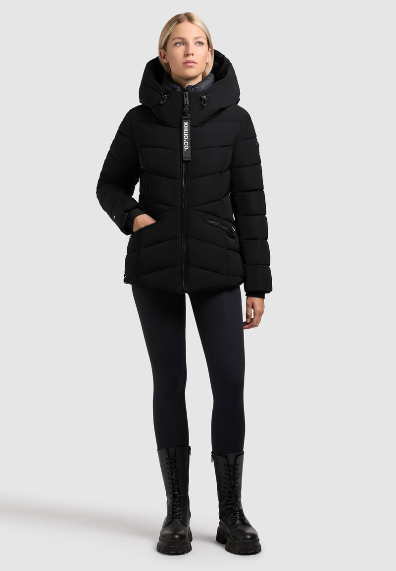 Jachetă puffer neagră cu glugă largă, închidere cu fermoar și cusături orizontale. Dispune de buzunare cu fermoar și un eticheta cu logo. Purtaa cu leggings negri și cizme.