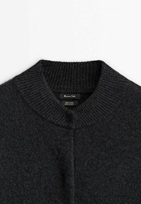 Donkergrijze wollen trui met een geribbelde kraag en een knooploze voorkant. Label zichtbaar met merk- en stofdetails. Zachte textuur.