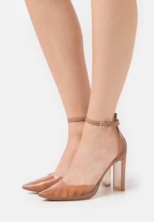 Läpinäkyvät, teräväkärkiset korkokengät, joissa on ruskea ankle strap ja kirkas lohkokanta, sekä sileä, kiiltävä pinta.