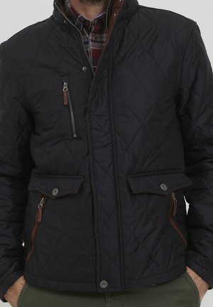 Uomo che indossa una giacca trapuntata nera con zip e tasche con bottoni sopra una camicia rossa a quadri, mani nelle tasche dei pantaloni verdi.