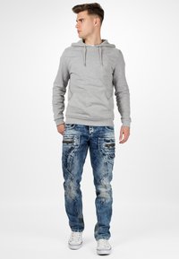 Felpa grigia con cappuccio e lacci, abbinata a jeans blu sbiaditi con tasche e dettagli di cucitura. Sneakers bianche completano l'outfit.