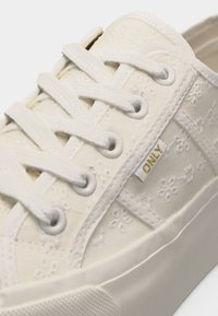 Baskets blanches avec broderie florale, lacets blancs, œillets métalliques, semelle en caoutchouc épaisse et une petite étiquette en tissu portant la mention "ONLY" sur le côté.