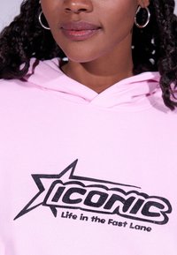 Lys rosa hettegenser laget av bomull, med en stor svart grafikk "Iconic" logo med en stjerne og teksten "Life in the Fast Lane" under.