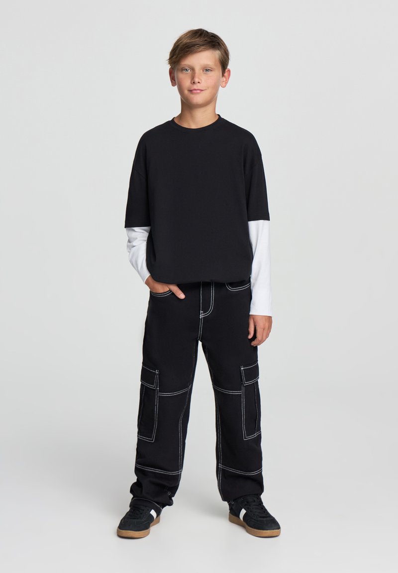 T-shirt nera a collo rotondo sovrapposta a una camicia bianca a maniche lunghe, abbinata a pantaloni cargo neri con cuciture bianche e tasche. Sneakers nere.