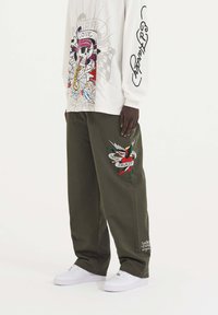 Witte blouse met lange mouwen met kleurrafgrafiek en tekst, gecombineerd met groene katoenen broek met geborduurde details en witte sneakers.