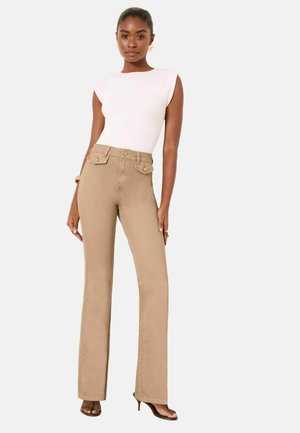 Woman wearing white sleeveless top, high-waisted beige pants with front flap pockets, and open-toe heels, standing against a plain white background.