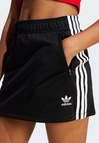 Črna športna krila iz mehke tkanine, z belimi stranskimi črtami, elastičnim pasom in žepom na zadrgo. Adidasov logotip na robu.