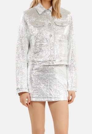 Femme portant une veste texturée argentée brillante avec des boutons et une mini-jupe assortie sur un fond uni.