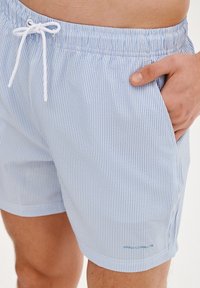 Pasiasty strój kąpielowy w jasnoniebieskim i białym kolorze, z elastycznym pasem w talji i sznurkiem do regulacji, kieszeniami bocznymi oraz małym detalem z logo.