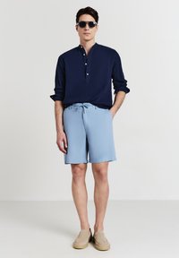 Dunkelblaues, strukturiertes Langarmhemd mit Knopfleiste, hellblaue Shorts mit Kordelzug und beiges Slipper. Modell trägt Sonnenbrille.