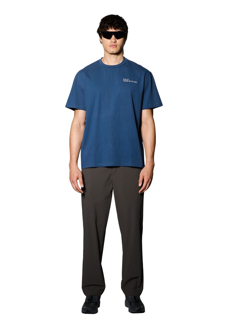 Blå kortärmad bomullströja med rund hals, med en liten logotyp på bröstet, tillsammans med bruna byxor och svarta skor.