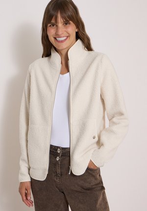 Femme souriante portant une veste polaire blanche zippée sur une chemise blanche et un pantalon marron taille haute avec deux boutons.
