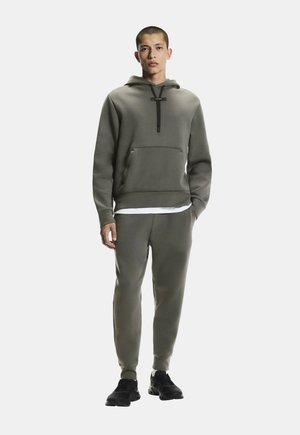 Jeune homme debout portant un sweat à capuche vert olive et un pantalon de survêtement assorti avec des baskets noires sur un fond blanc uni.