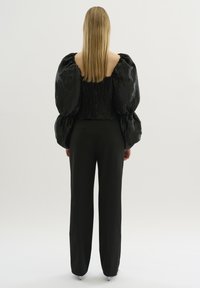 Zwarte textuurtop met volumineuze, opbollende mouwen, gecombineerd met zwarte broek. Glad materiaal en geplooide details accentueren het ontwerp.