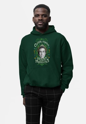 Man in een donkergroene hoodie met een afbeelding en de tekst "Ik ben een Cotton-Headed Ninny Muggins" en zwarte broek met witte vierkantjes.